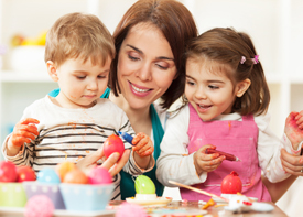 Eine Erzieherin sitzt mit zwei kleinen Kindern an einem Tisch. Sie bemalen Ostereier.