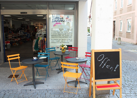 Am Eingang von Tonis Ladencafé stehen drei kleine Kaffee-Tische mit mehreren Stühlen. Auf einem Klappschild steht "Offen!"