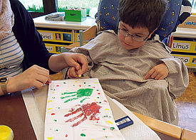 Ein kleiner Junge im Rollstuhl malt mit Hilfe einer Assistenzkraft ein Bild.