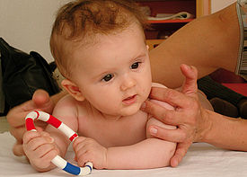 Ein Baby liegt auf dem Bauch und hält einen Greifring, zwei Hände einer Förderkraft stützen es.