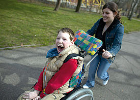 Ein Junge im Rollstuhl wird von einer Assistenzkraft durch einen Park geschoben.