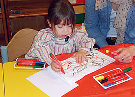 Ein kleines Mädchen malt mit Buntstiften. Neben ihr sitzt eine Schulbegleiterin.