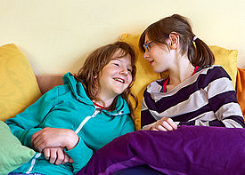 Zwei Mädchen liegen gemütlich auf einem Sofa und unterhalten sich.