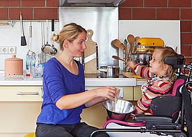 Ein Mädchen im Rollstuhl sitzt mit einer Assistenzkraft in einer Küche. Sie rühren zusammen in einer Schüssel.