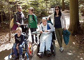 Zwei Kinder im Rollstuhl stehen im Wald zusammen mit zwei Betreuern und einem weiteren Jungen.