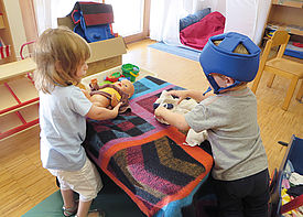 Zwei kleine Kinder mit Behinderung stehen an einem Tisch und wickeln eine Puppe.