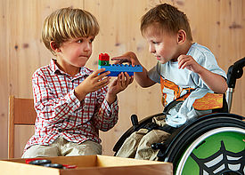 Zwei kleine Jungs bauen zusammen mit Duplo-Steinen. Einer von ihnen sitzt im Rollstuhl.