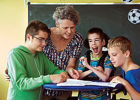 Drei Jungs mit Behinderung und eine Lehrerin sind im Klassenzimmer. Sie stehen an einem Tisch und beugen sich über ein Heft.