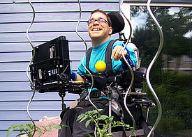 Ein erwachsener Mann im Rollstuhl steht im Garten neben einem Haus und lacht.