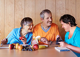 Drei erwachsene Menschen mit Behinderung sitzen an einem Tisch und schneiden Obst.