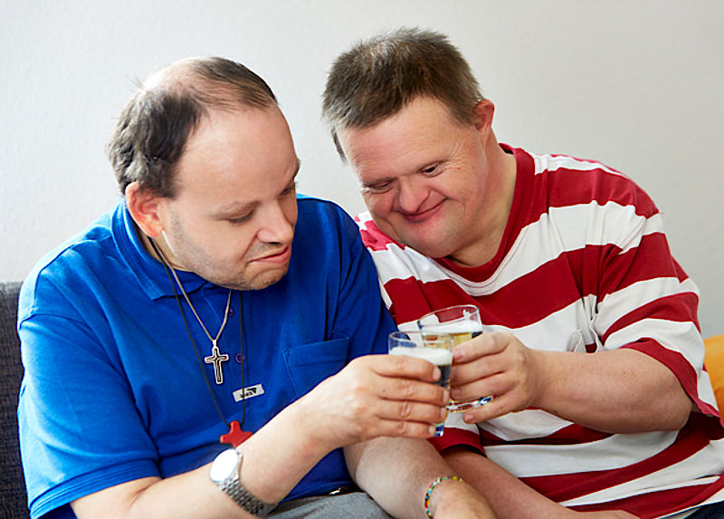 Zwei Männer mit Behinderung sitzen auf einem Sofa und stoßen gemeinsam an.
