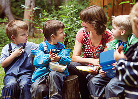 Vier Kinder und eine Erzieherin sitzen im Wald und vespern.