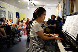 Eine junge Frau spielt auf dem Klavier in einer Runde von Zuhörern.