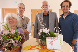 Vier Personen blicken lächelnd in die Kamera. Die Person ganz links hält einen Blumenstrauß.