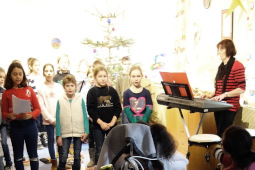 Eine Gruppe Schüler steht vor einem Weihnachtsbaum und singt. Links im Bild steht eine Lehrerin am E-Piano.