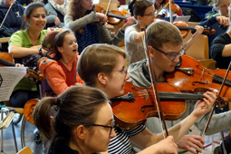 Ein Kind im Rollstuhl und eine Betreuerin sitzen mitten zwischen Jugendlichen, die Geige spielen.