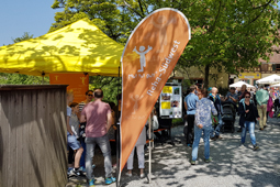 Viele Menschen laufen über einen Platz mit schattenspendenden Bäumen. Im Vordergrund sieht man einen Stand der Reha-Südwest.