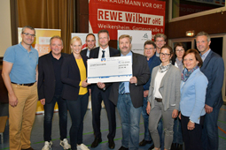 Zwölf Personen stehen in einer Halle und lächeln in die Kamera. Die drei Personen in der Mitte der Gruppe halten einen Spendenscheck in die Höhe.