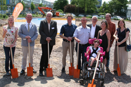 Auf dem Bild sind neun Erwachsene und ein Kind im Rollstuhl zu sehen. Sie stehen auf einer Baustelle, im Hintergrund ist ein Bagger zu sehen. Jeder hält einen Spaten in der Hand.