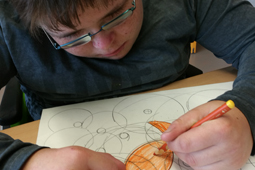 Ein Junge mit einer Brille sitzt vor einem Blatt Papier. Er zeichnet einen Fisch.