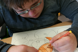 Ein Junge mit einer Brille sitzt vor einem Blatt Papier. Er zeichnet einen Fisch.