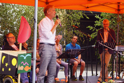 Ein Mann in einem feinen Hemd steht auf einer Bühne und spricht in ein Mikrofon. Im Hintergrund sind Leute an Instrumenten zu sehen.