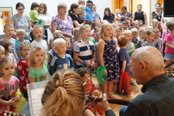 Eine große Gruppe Kinder und einige Erwachsene singen ein Lied. Im Vordergrund ist ein Mann an einer Gitarre zu sehen.