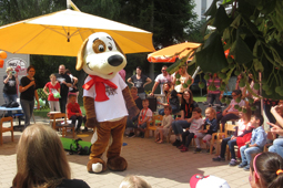 Ein als Plüschhund verkleideter Mensch steht auf einem Platz. Um ihn herum sitzen und stehen viele Kinder und Erwachsene.