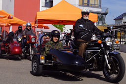 Ein Motorradfahrer und ein Beiwagenfahrer auf einem Schulhof. Im Hintergrund sind weitere Fahrer, Schirme und ein Fest-Pavillon zu sehen.