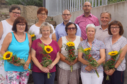 Zehn Erwachsene blicken in die Kamera. Einige von ihnen haben einen Strauß mit einer Sonnenblume in der Hand.