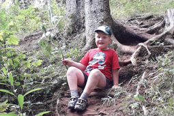 Ein Junge in kurzer Hose und Schirmmütze sitzt im Wald unter einem Baum.