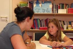 Eine Schulbegleiterin und ein Kind sitzen vor einem Bücherregal und schauen sich an.