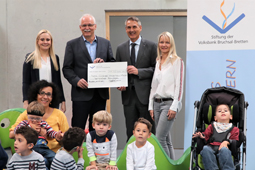 Vier Erwachsene stehen vor einer Wand mit einem Scheck in der Hand. Davor sitzen Kinder.