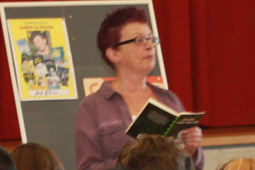 Eine Frau mit Brille steht vor einer Tafel und liest aus einem Buch.