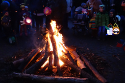 Im Vordergrund brennt ein Lagerfeuer, im Hintergrund sind Kinder mit Laternen zu sehen.