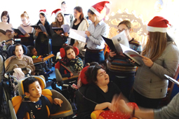 Mehrere Jugendlichen mit Nikolausmützen auf dem Kopf stehen und singen. Um sie herum sitzen Kinder in Rollstühlen.