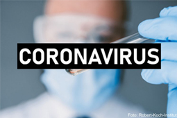 Ein Forscher mit Mundschutz und Schutzbrille hält ein Reagenzglas in die Kamera. Ein schwarzer Balken mit dem Schriftzug "Coronavirus" verdeckt das Bild teilweise.