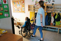 Eine Freiwillige schiebt ein Kind im Rollstuhl durch den Kindergarten, vorbei an einer Garderobe.