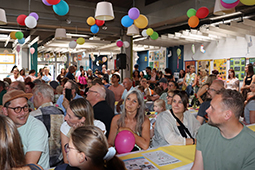 Besucher des Sommerfests der Kita Regenbogen an Tischen sitzend. Bunte Luftballons an der Decke.