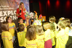 Eine Gruppe Kinder steht an einem Stand und sieht einem Kind zu, das ein gebasteltes Kunstwerk präsentiert.