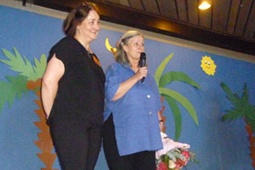 Zwei Frauen stehen auf einer Bühne. Eine hält einen Blumenstrauß in der Hand und spricht in ein Mikrofon.