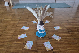 Ein Yogaraum, in dem in der Mitte eine Vase mit Trockenblumen und einer weiblichen Puppe steht. Drumherum liegen 8 Karten im Kreis. Im Hintergrund liegt eine blaue Yogamatte.